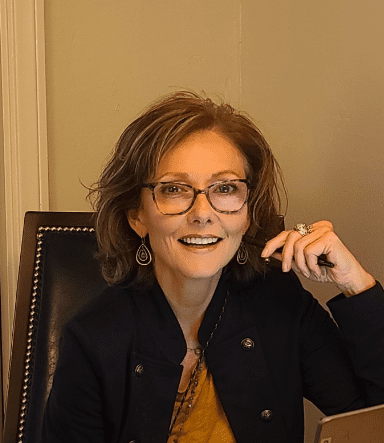 484586030_614212951614846_4688559580962504365_n Person with short brown hair and glasses, smiling, seated in a dark chair with studded backrest, wearing a dark jacket and earrings, holding a pen.