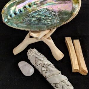 An abalone shell, white crystal, White Sage Smudging kit, and pieces of palo santo wood arranged on a black background.