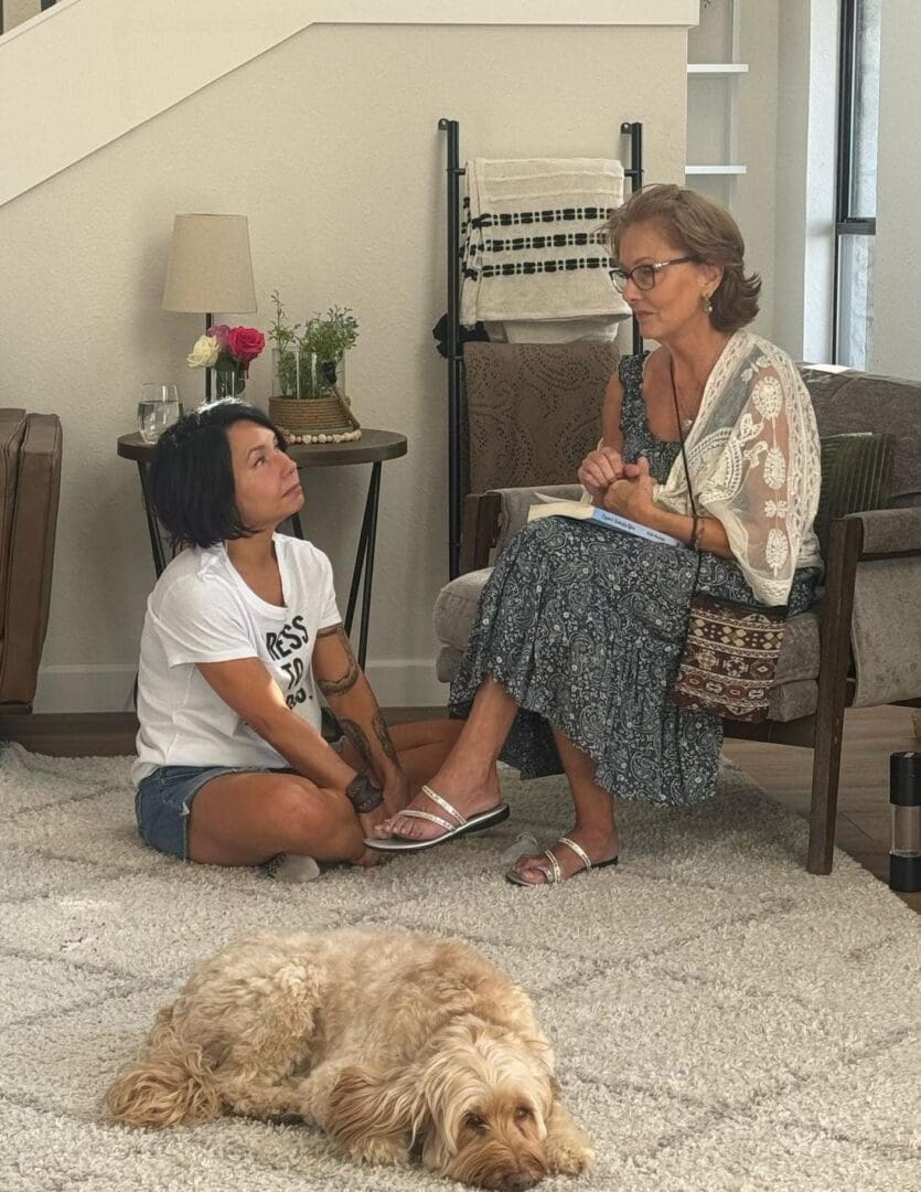Two women have a conversation in a living room; one sits on a chair holding a notebook while the other sits cross-legged on the floor. A dog lies on the rug in the foreground.