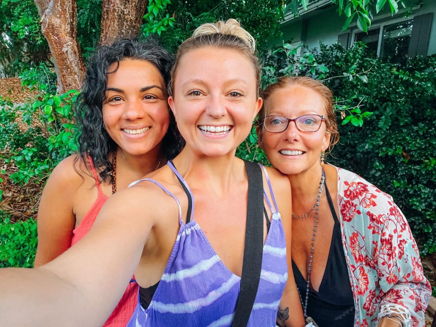 Three women stand close together outdoors, smiling at the camera. Green foliage and part of a building are visible in the background.