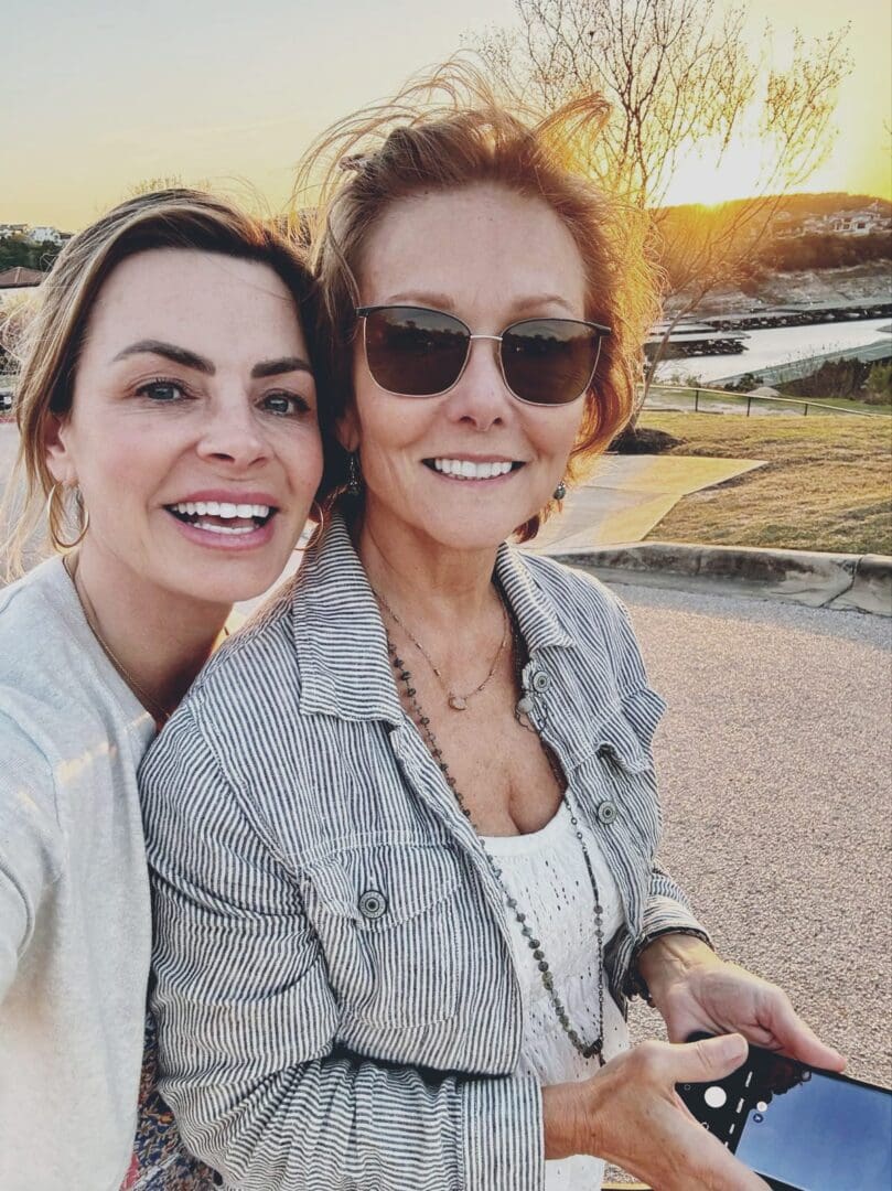 Two women smiling outdoors at sunset, one holding a smartphone, with a body of water and trees in the background.
