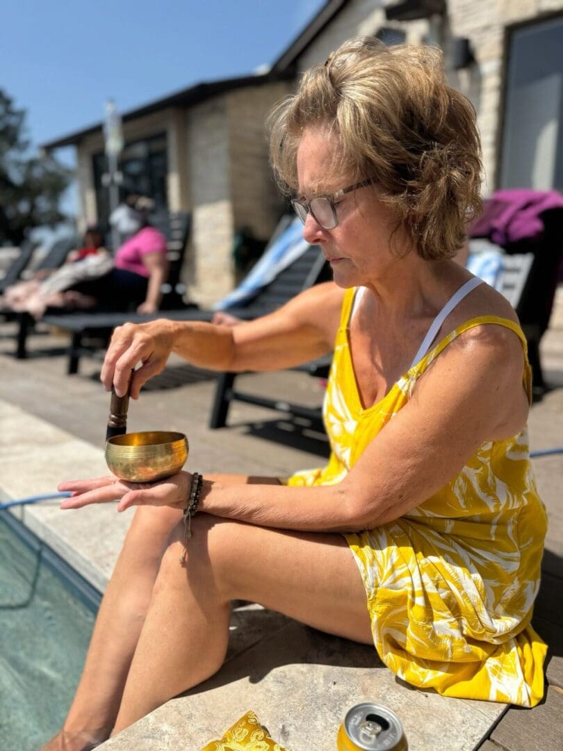 A woman in a yellow dress sits by a pool, using a mallet on a singing bowl balanced on her hand. A can is on the ground nearby, and people lounge in the background.