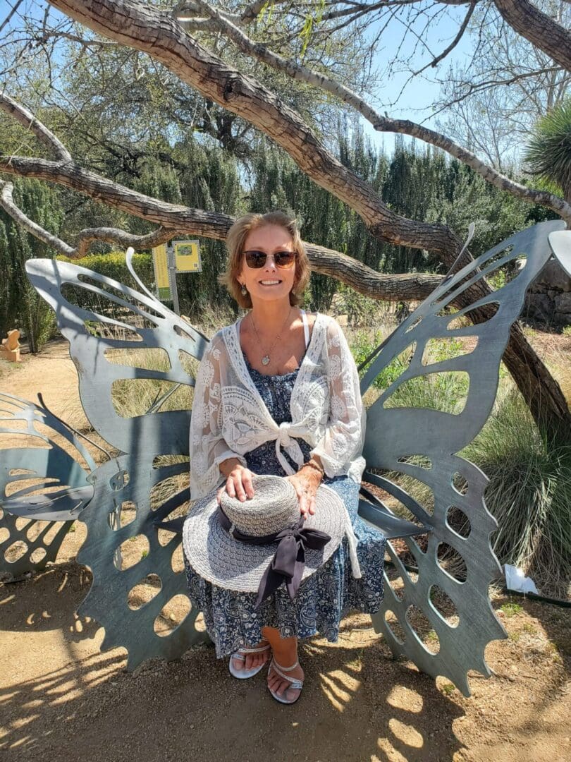 Woman sitting on a butterfly-shaped metal bench outdoors, wearing sunglasses, a lace cover-up, and holding a hat on her lap. Trees and greenery surround the scene.