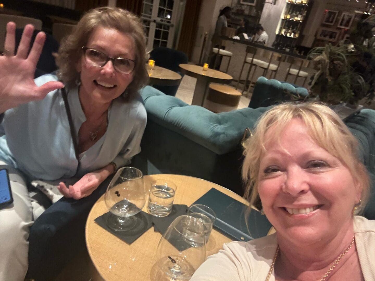 Two women sit at a round table in a lounge, smiling at the camera. One is waving; there are empty glasses and water glasses on the table.