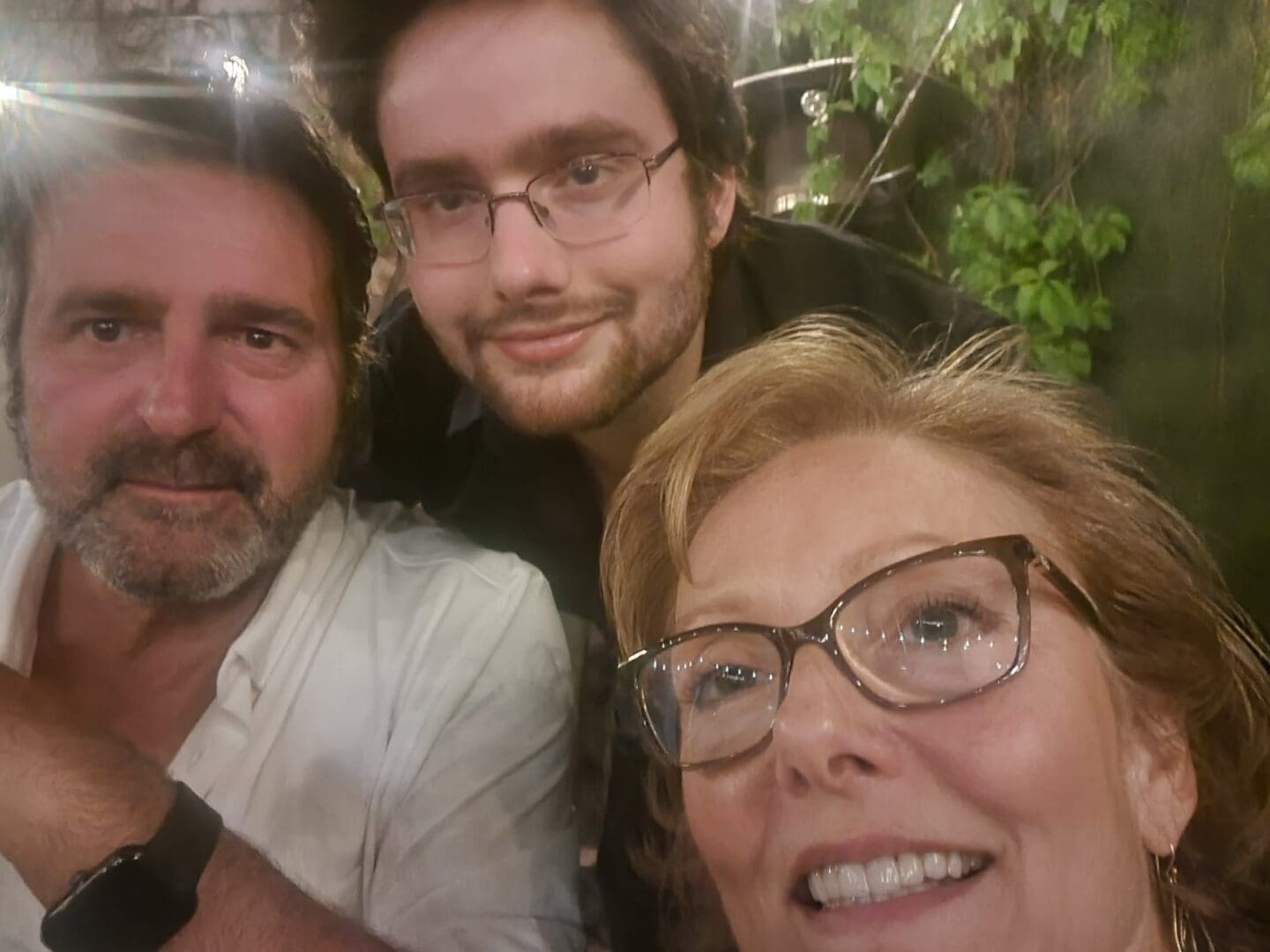 Three people pose closely together for a selfie, all wearing glasses, with greenery and outdoor lighting visible in the background.
